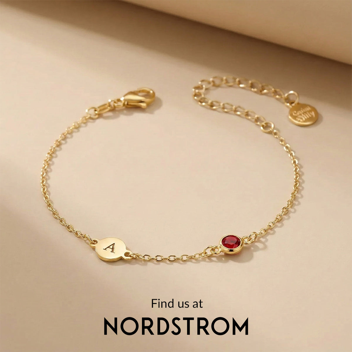 Gold bracelet with a red gemstone and 'A' charm on a beige background, Nordstrom branding visible.