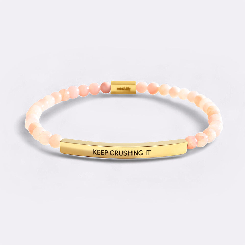 Bracelet with pink beads and gold bar with 'KEEP CRUSHING IT' text on a light gray background