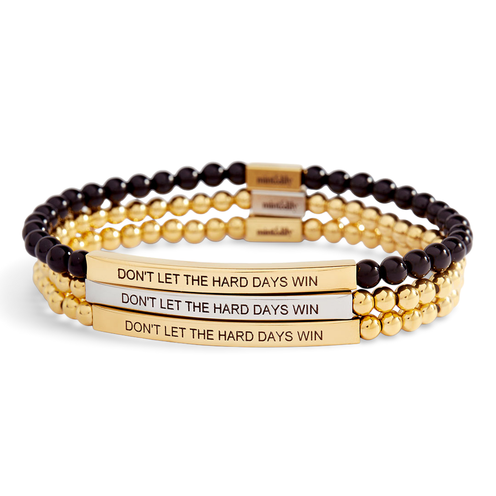 Stack of gold and black beaded bracelets with motivational message on a light gray background