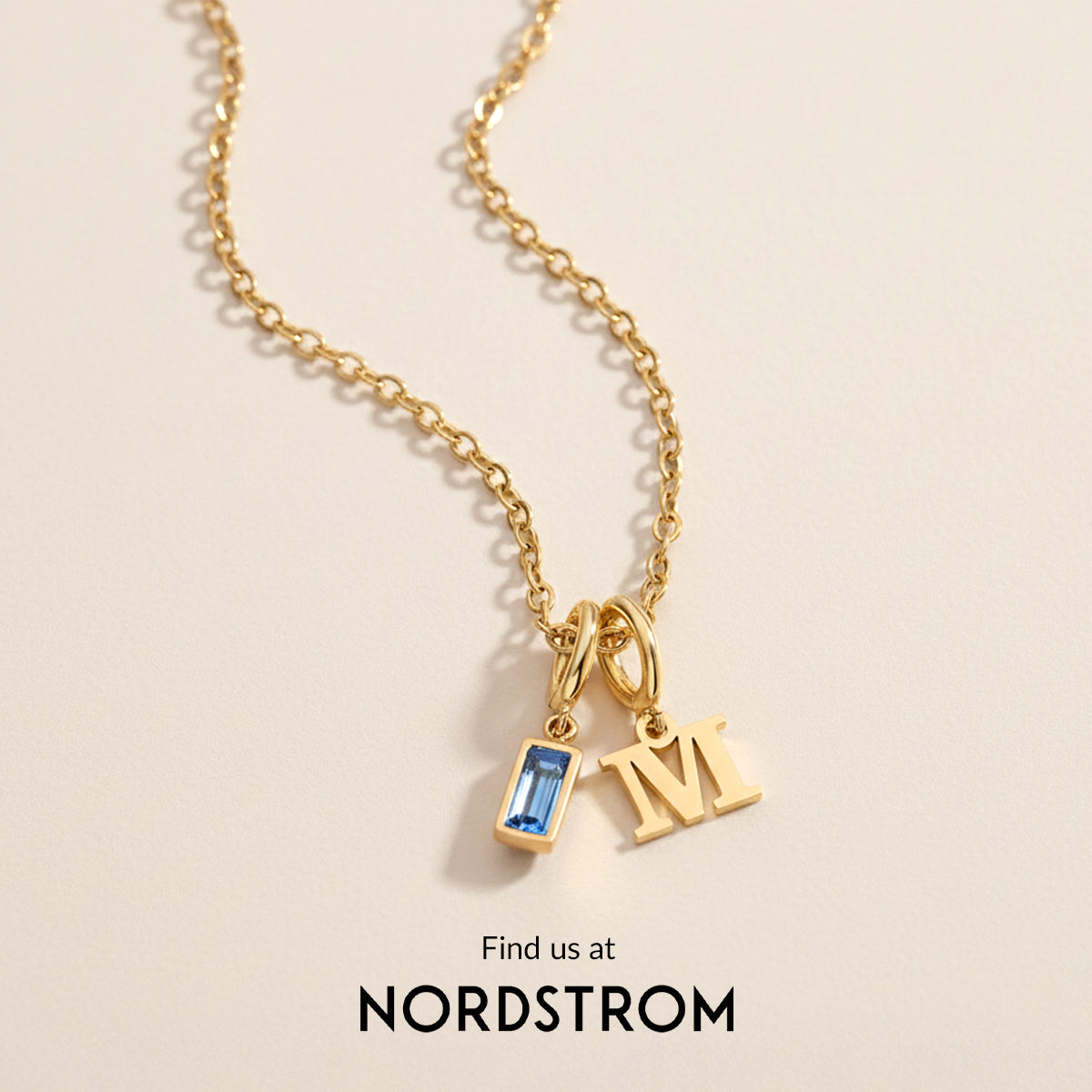Gold necklace with 'M' pendant and blue gemstone on a beige background, Nordstrom branding.