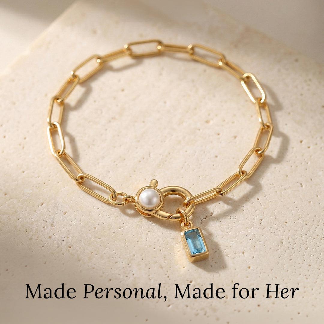 Gold chain bracelet with a pearl and blue gemstone charm on a beige background.