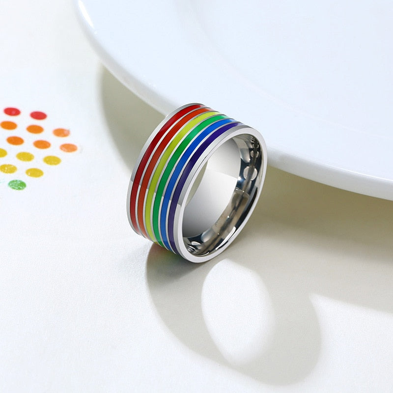 A stainless steel band ring with a rainbow-colored enamel stripe, placed on a white surface next to a white plate with colorful polka dots.