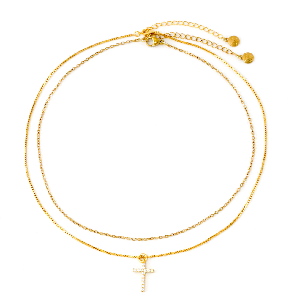 Gold dainty cable necklace and gold pavé cross chain necklace laid flat in concentric circles on a white background, clasps and extenders visible.