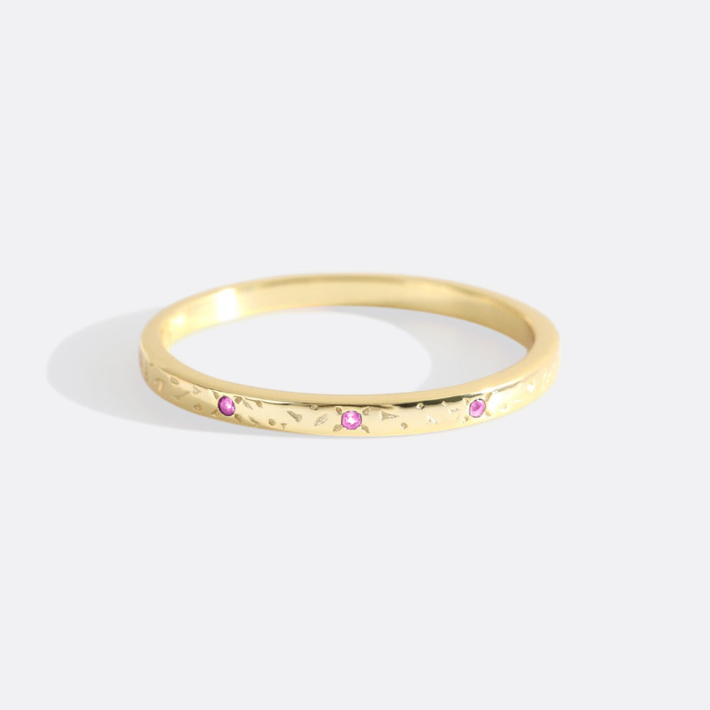 A gold-colored narrow band ring with textured surface and small red gemstones set on it.